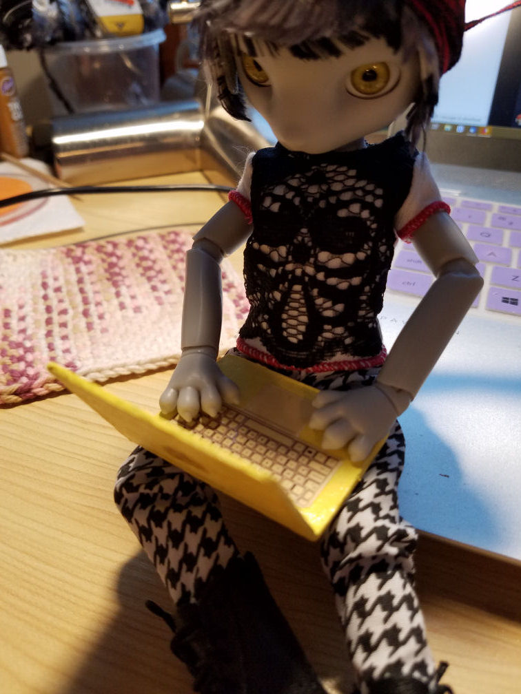 A grey Hujoo Phoebe Mouse doll sitting on the edge of a silver laptop with a doll sized pale yellow laptop open on her lap. Looking over the laptop lid we can see the yellow keyboard.